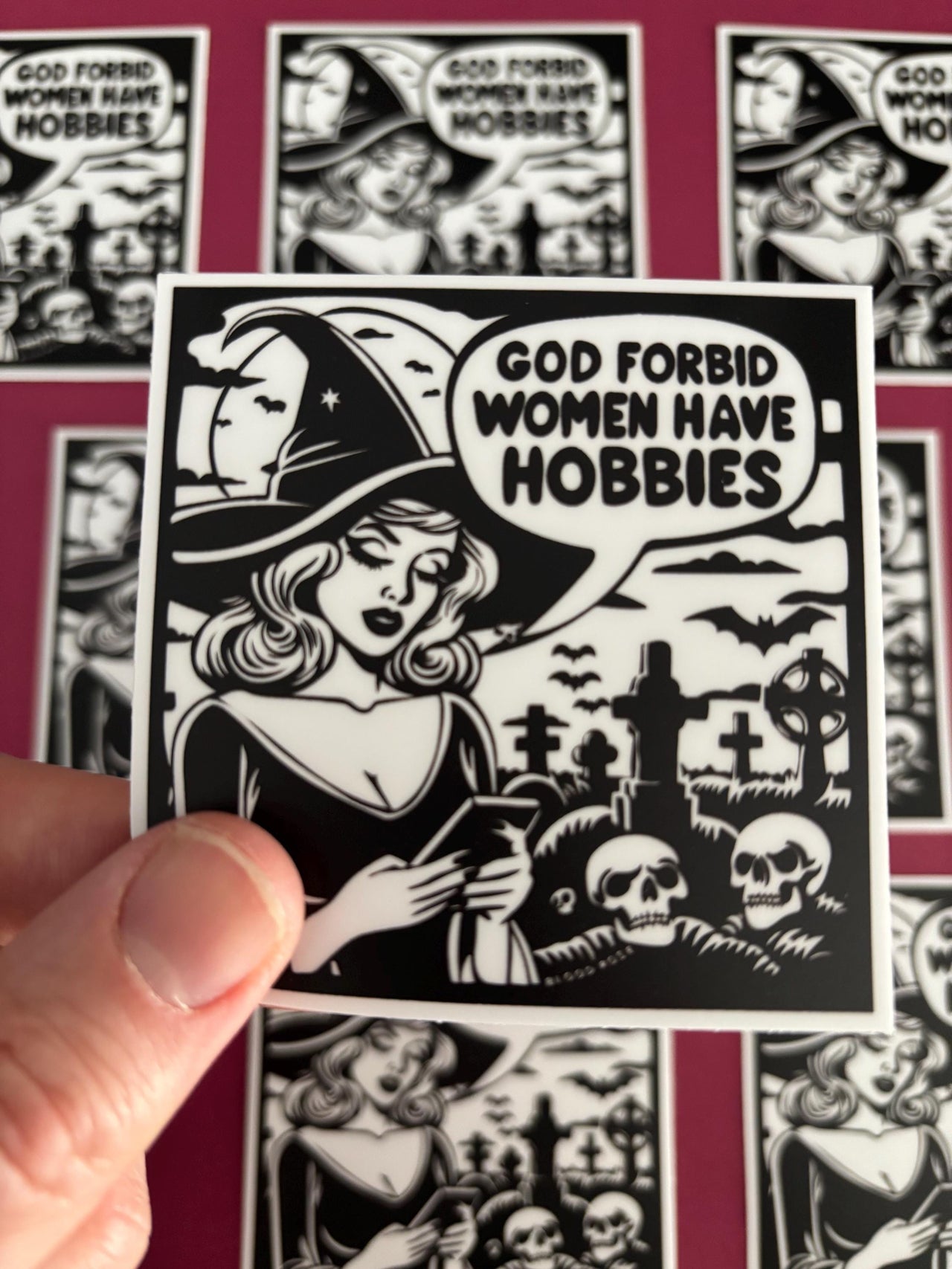 Sticker with a woman in a witch hat and text 'God Forbid Women Have Hobbies' held by a hand against a background of similar stickers.